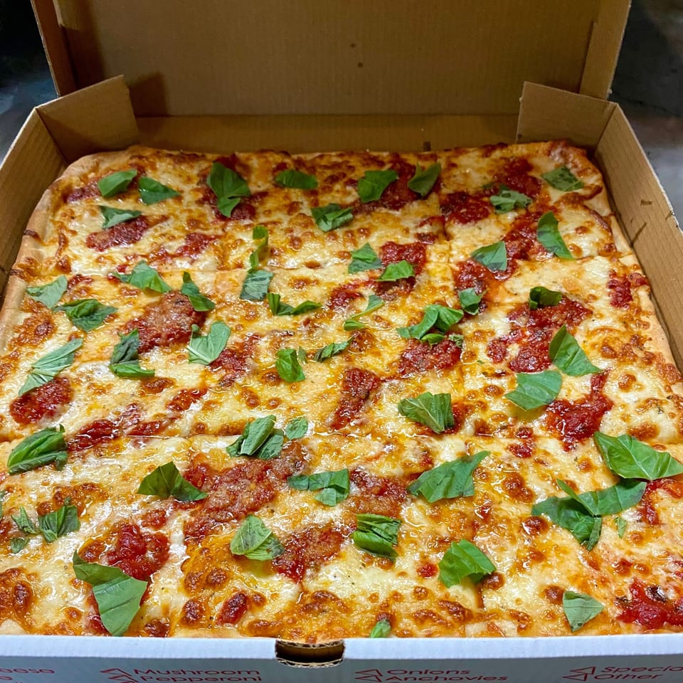 A square Capri Pizza with melted cheese, red sauce, and fresh basil leaves in a cardboard box.