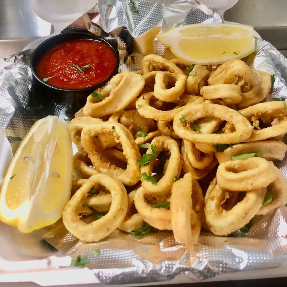 Crispy fried calamari rings with marinara sauce and lemon wedges, served in a foil-lined container.