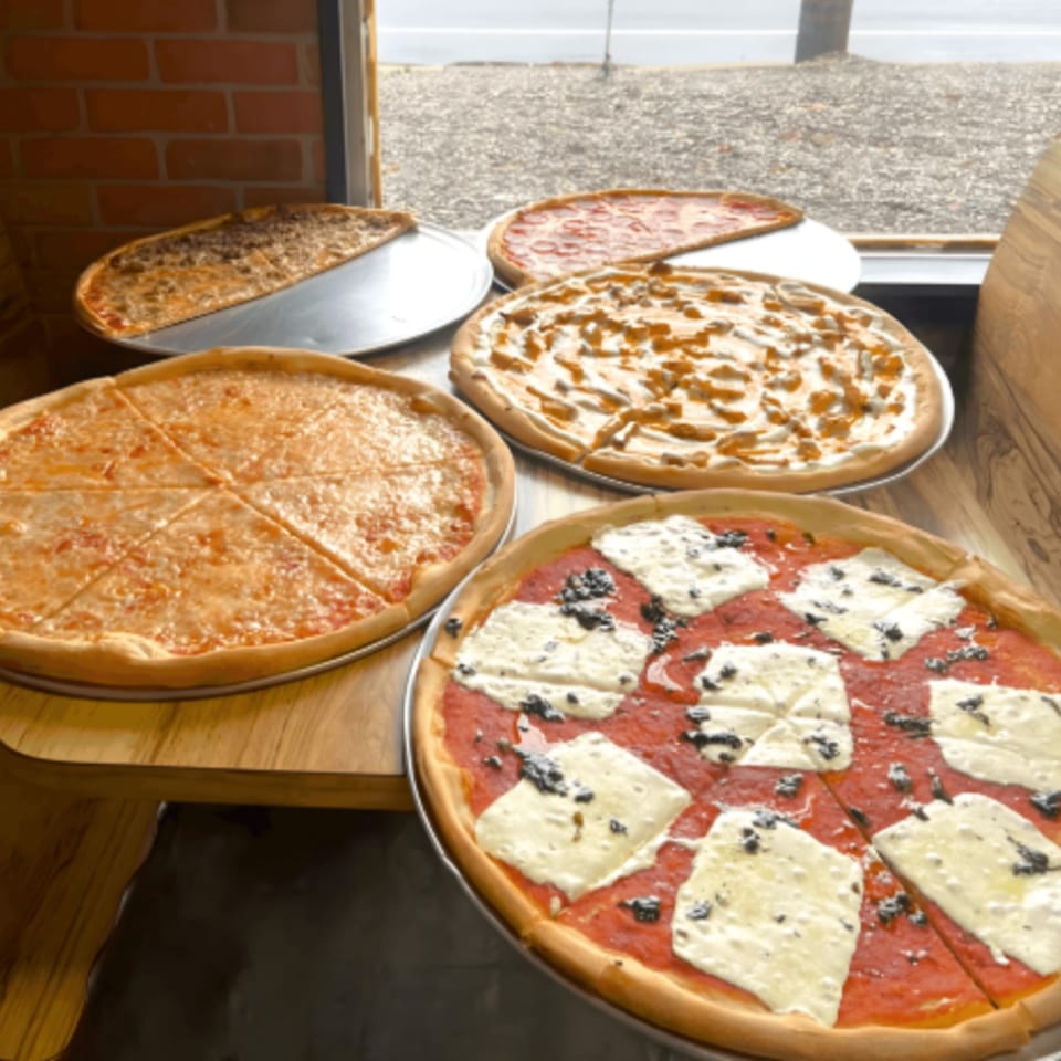 Multiple Capri Pizza pizzas, including cheese, pepperoni, and specialty pies, on a wooden table.