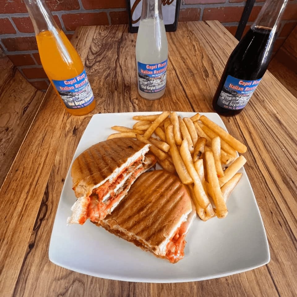 A Capri Pizza panini with fries and three Capri Pizza branded sodas on a wooden table.