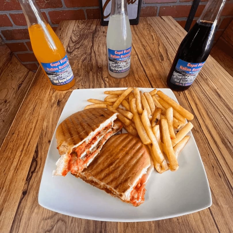 A Capri Pizza panini with fries and three Capri Pizza branded sodas on a wooden table.